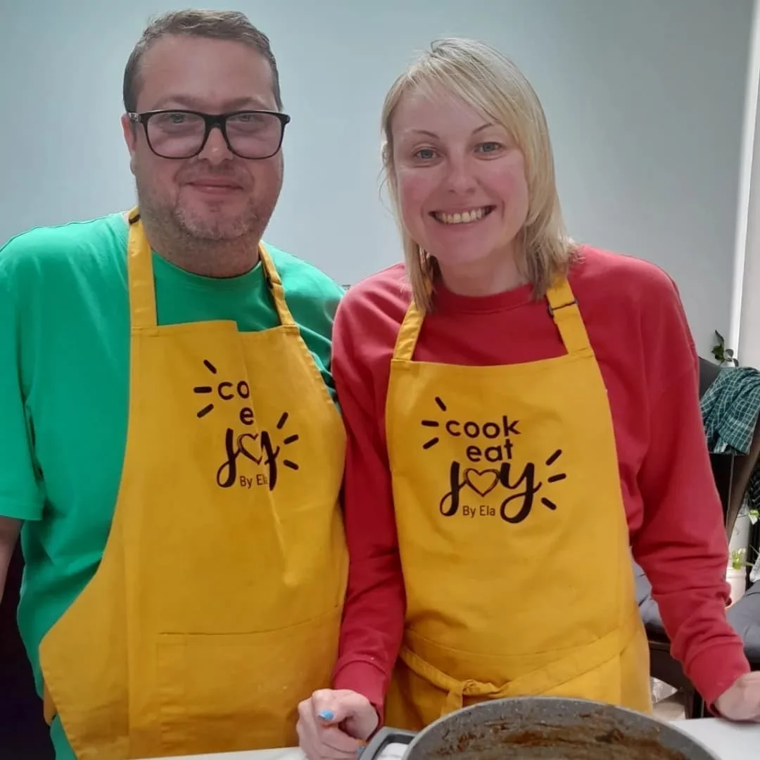 Couple cooking together in Cook Eat Joy yellow aprons