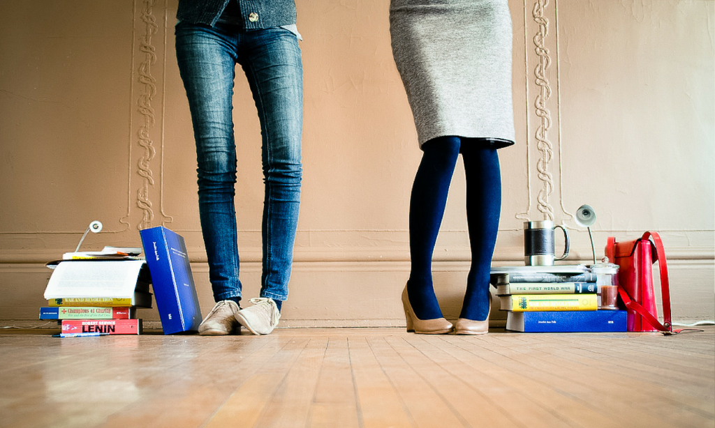 Two people standing against a panelled wall, surrounded by stacks of academic books at their feet.