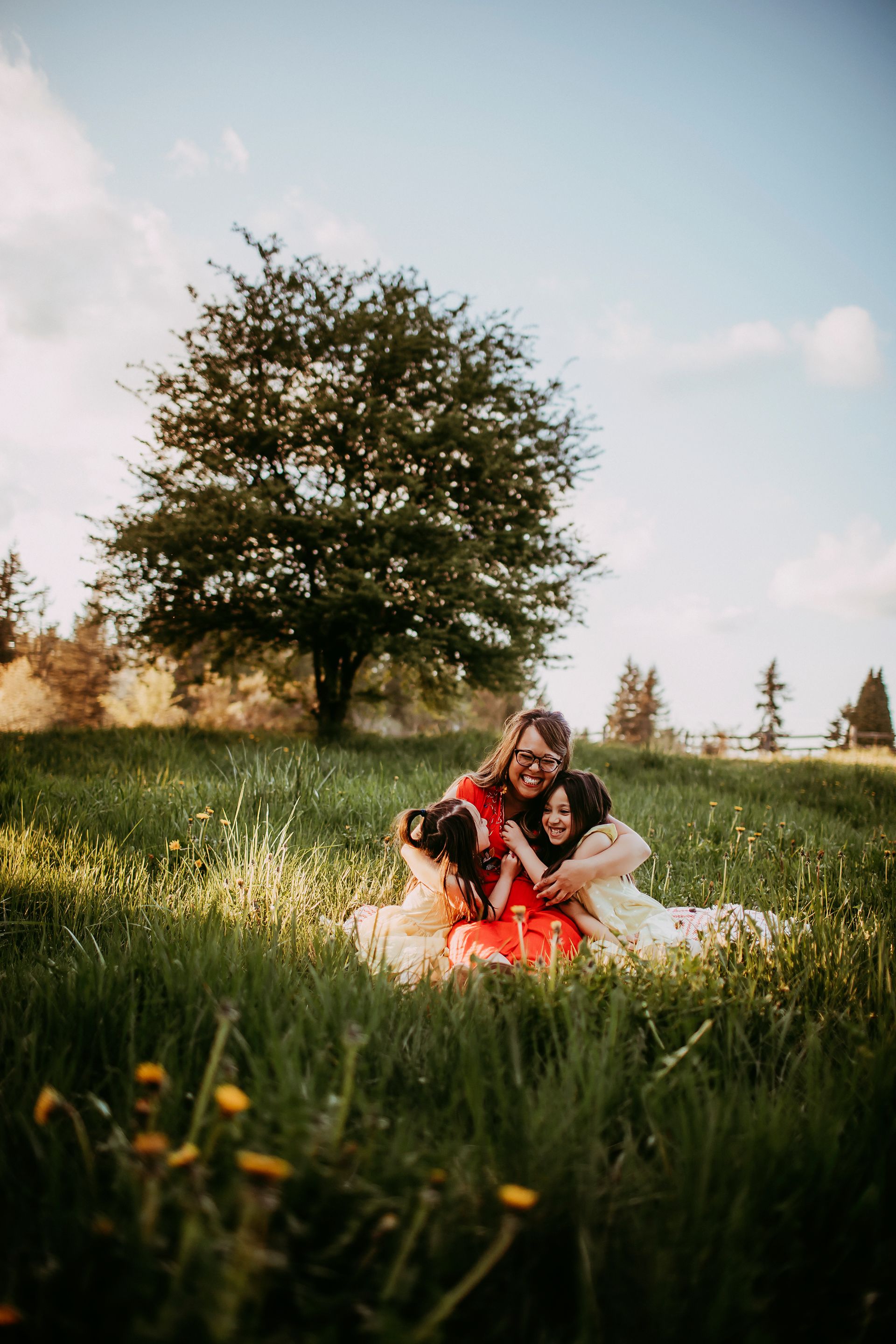 Mona sitting in a sunlit meadow, laughing with her two daughters wrapped around her.