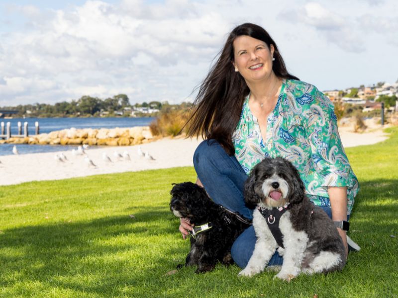 Carolyn Verhoef sitting on the grass by the Swan River with her two dogs.